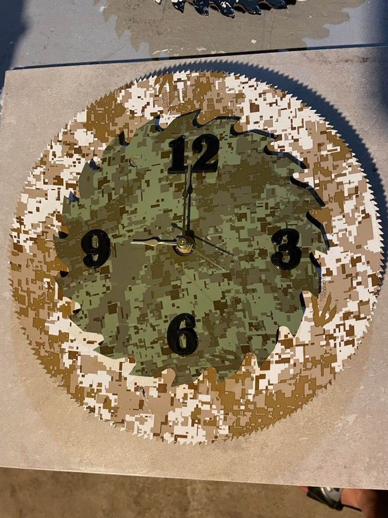 A circular wall clock made from a saw blade with a camouflage pattern and black numbers 3, 6, 9, and 12.