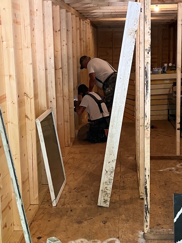 Two people in work clothes install structural components inside a wooden frame building under construction.
