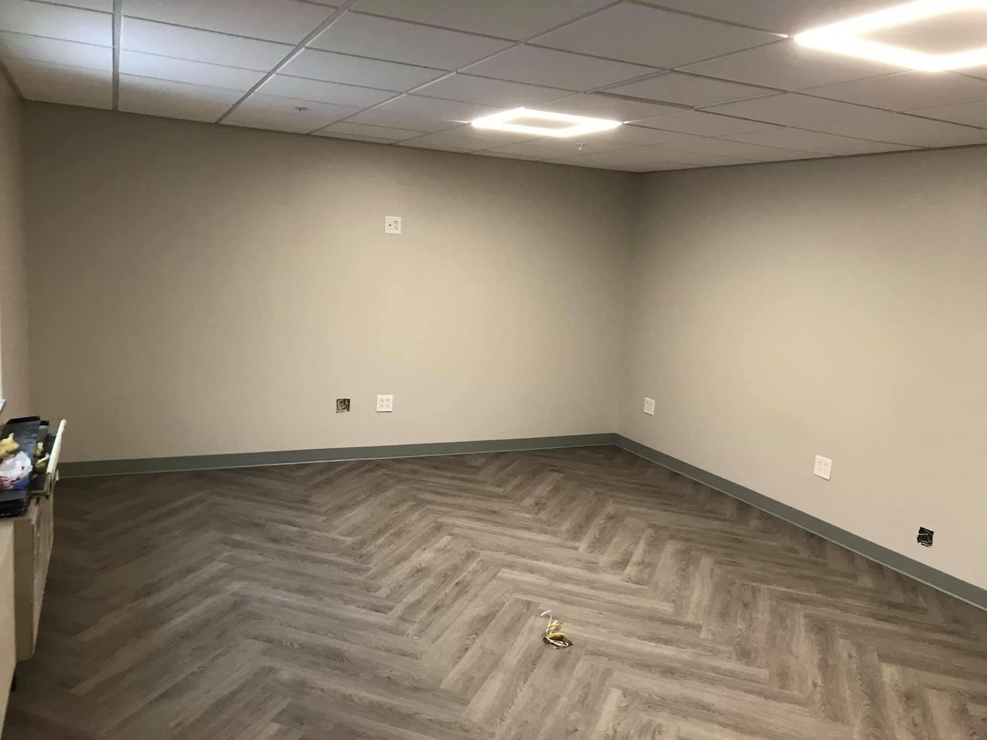 An empty, modern office space with light gray walls, dark wood-look herringbone flooring, and recessed ceiling lights.