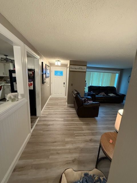 A hallway leads to a white front door, with a living room featuring dark leather furniture to the right.