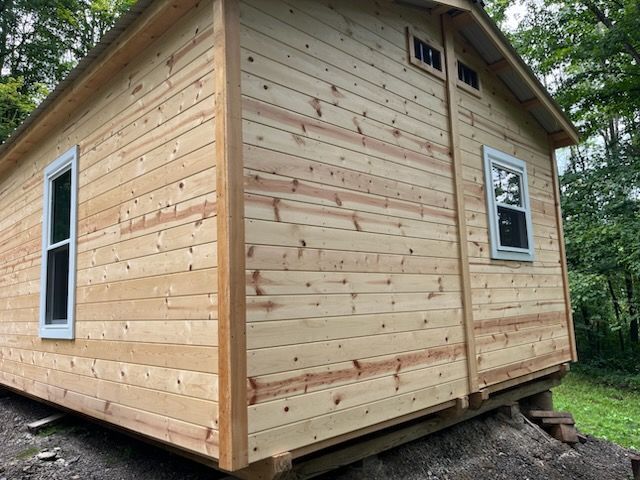 A side view of a small, light-colored wooden cabin on a foundation, featuring two windows and wood panel siding.