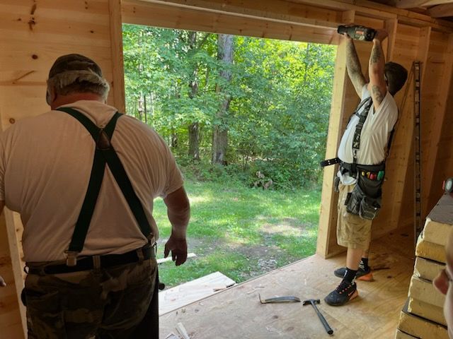 Two construction workers in tool belts frame a large doorway in a wooden structure overlooking a wooded area.