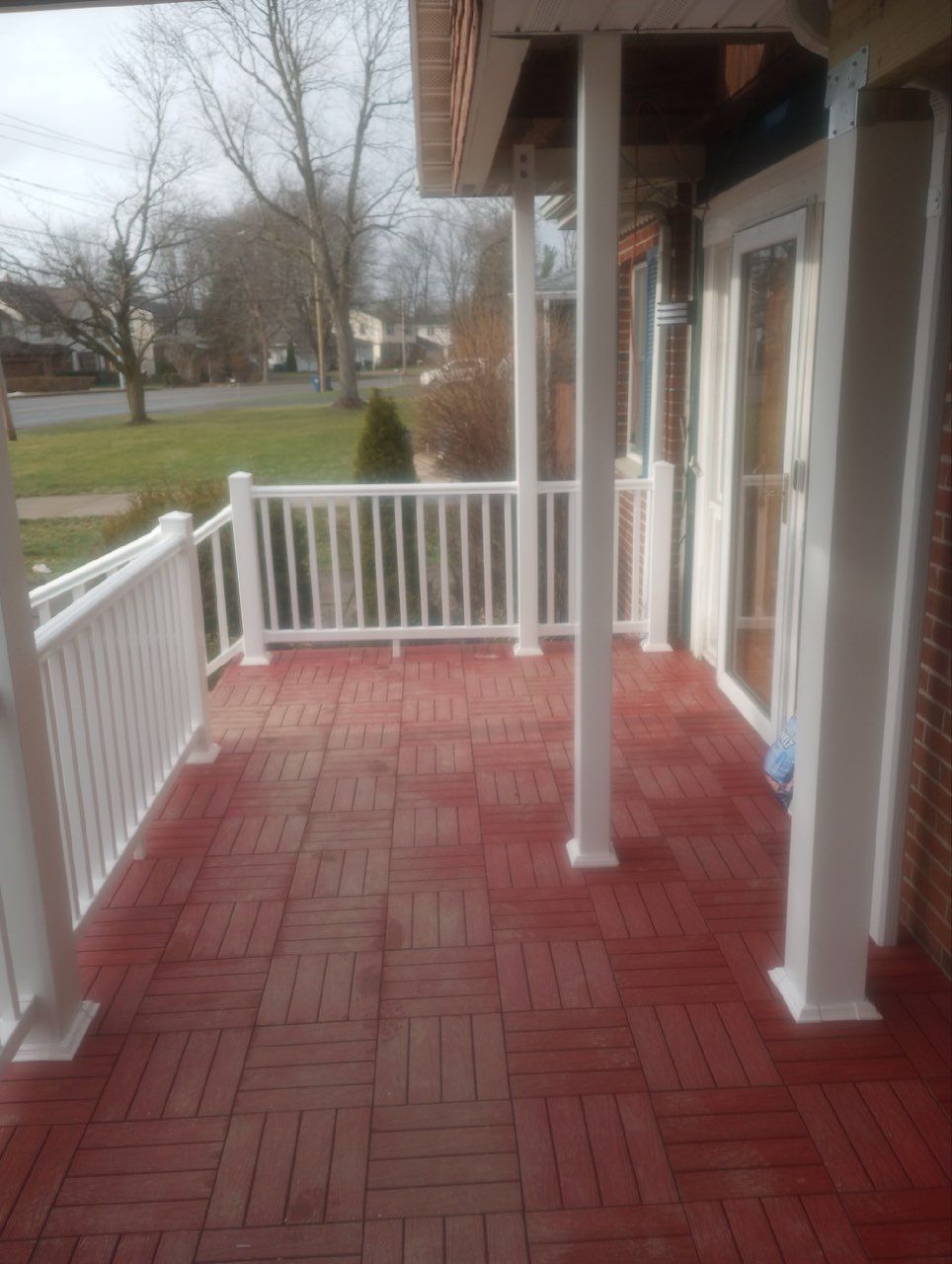A residential porch with reddish-brown tiled flooring, white railings, and white support posts under a roof.
