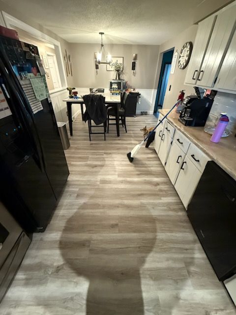 A kitchen and dining area with white cabinets, a black refrigerator, wood-look flooring, and a mop leaning on the counter.