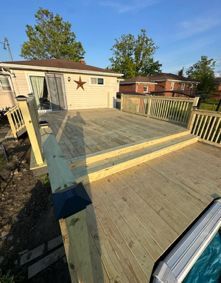 A tiered wooden deck with railings extends from a house toward a backyard pool on a sunny day.