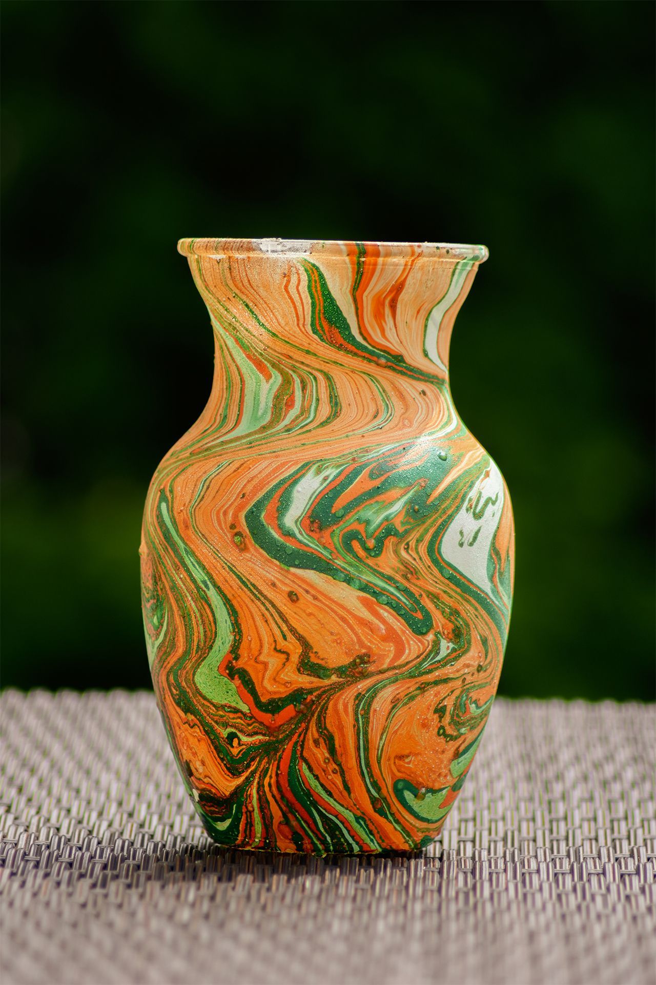 A small vase with a marbled, swirl pattern of orange, green, and cream, resting on a textured, dark brown surface.
