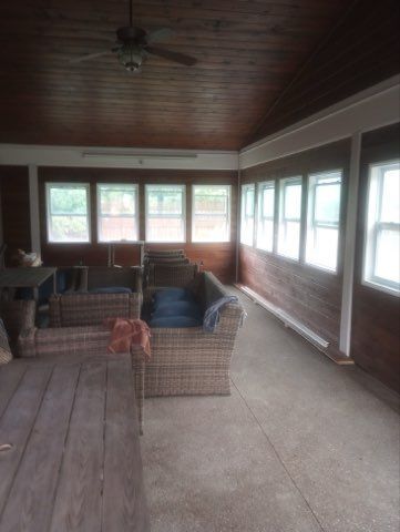 A rustic, enclosed sunroom with wooden walls, a vaulted ceiling with a fan, wicker furniture, and large windows.