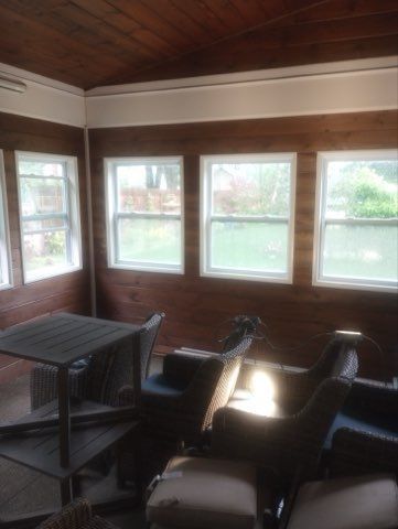 A sunroom with wood-paneled walls and four large windows, featuring a table and several chairs with cushions.