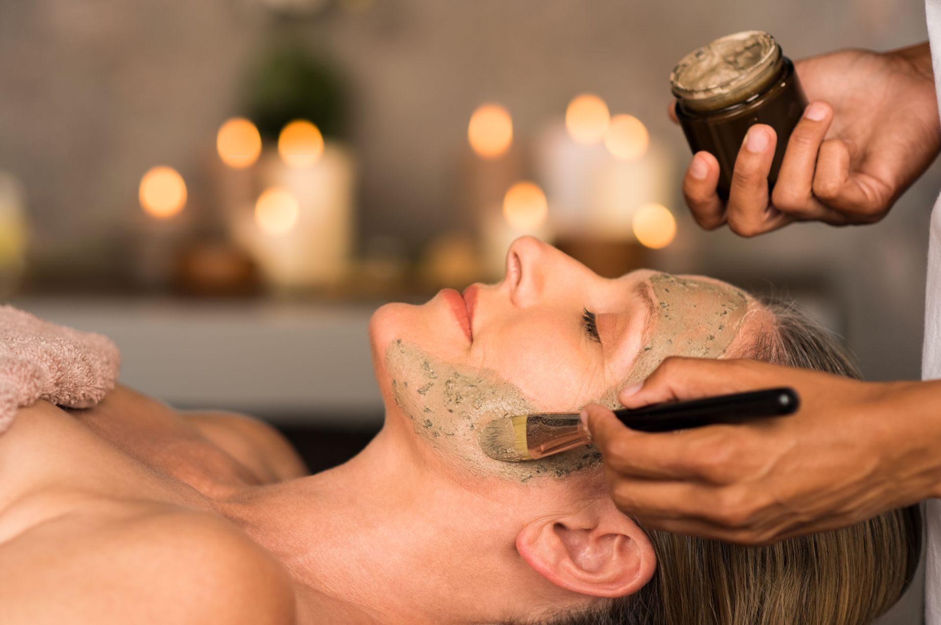Gesichtsbehandlung im Spa: Einer liegenden Kundin wird Creme aufgetragen, während im Hintergrund Kerzen leuchten