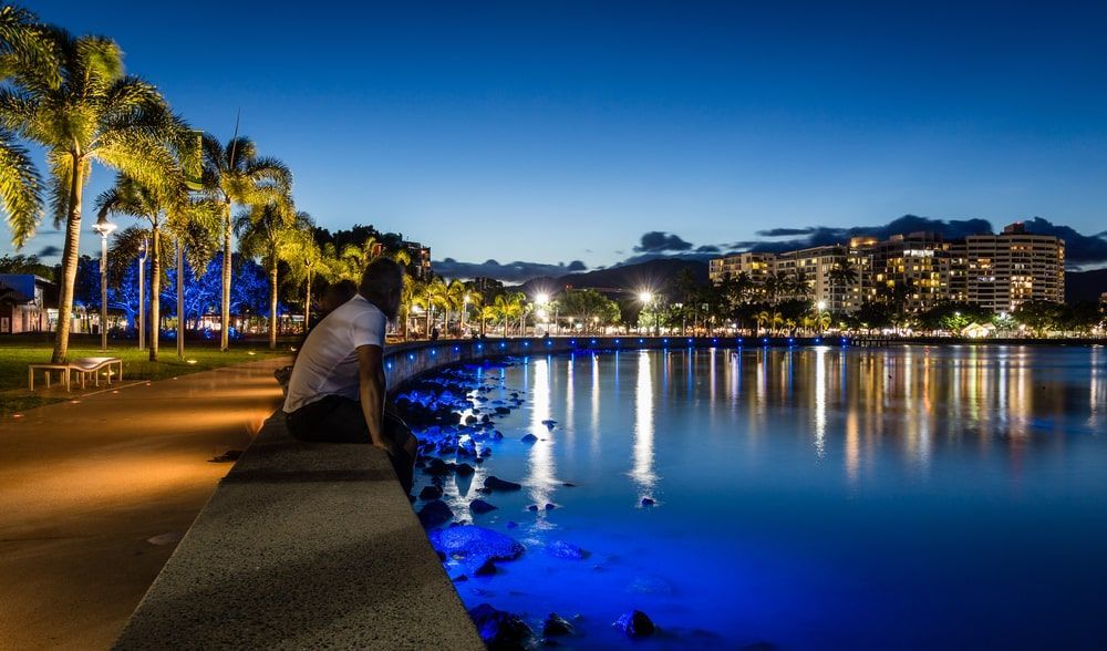 A Man Is Sitting On The Edge Of A Body Of Water At Night — Lux Noir In Cairns, QLD