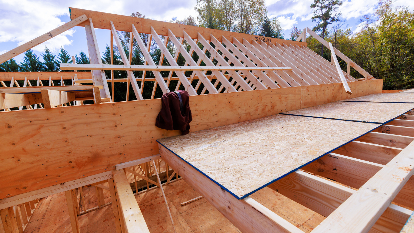 Wooden roof construction in progress; trusses, plywood, and lumber visible outdoors.