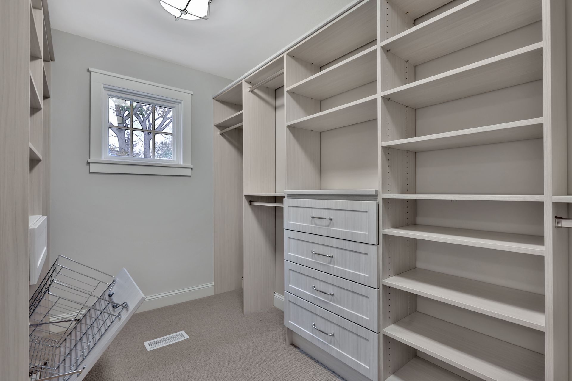 Empty walk-in closet with white built-in shelves, drawers, and a small window, carpeted floor.