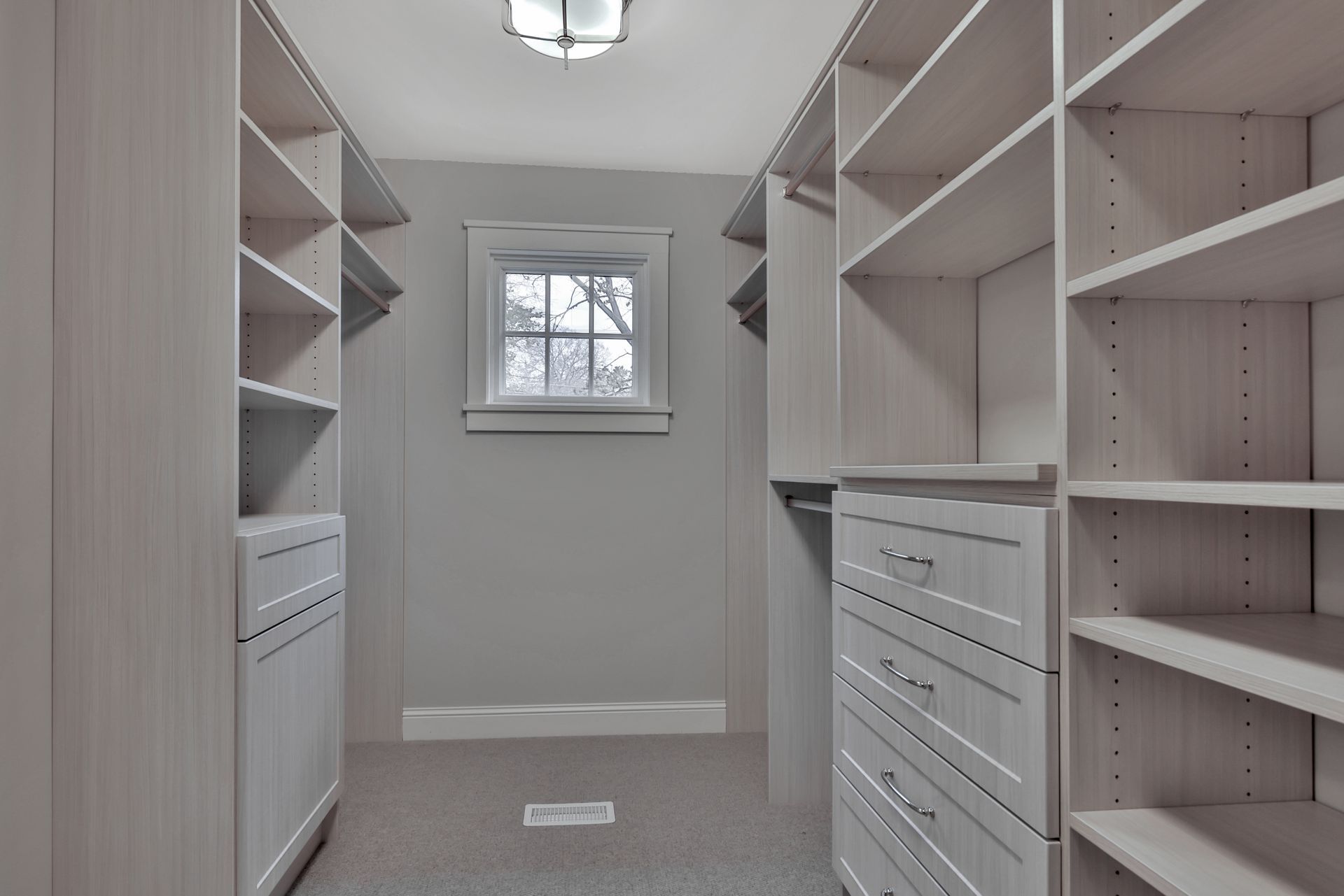 Empty walk-in closet with white built-in shelves and drawers, beige carpet, and a small window at the end.