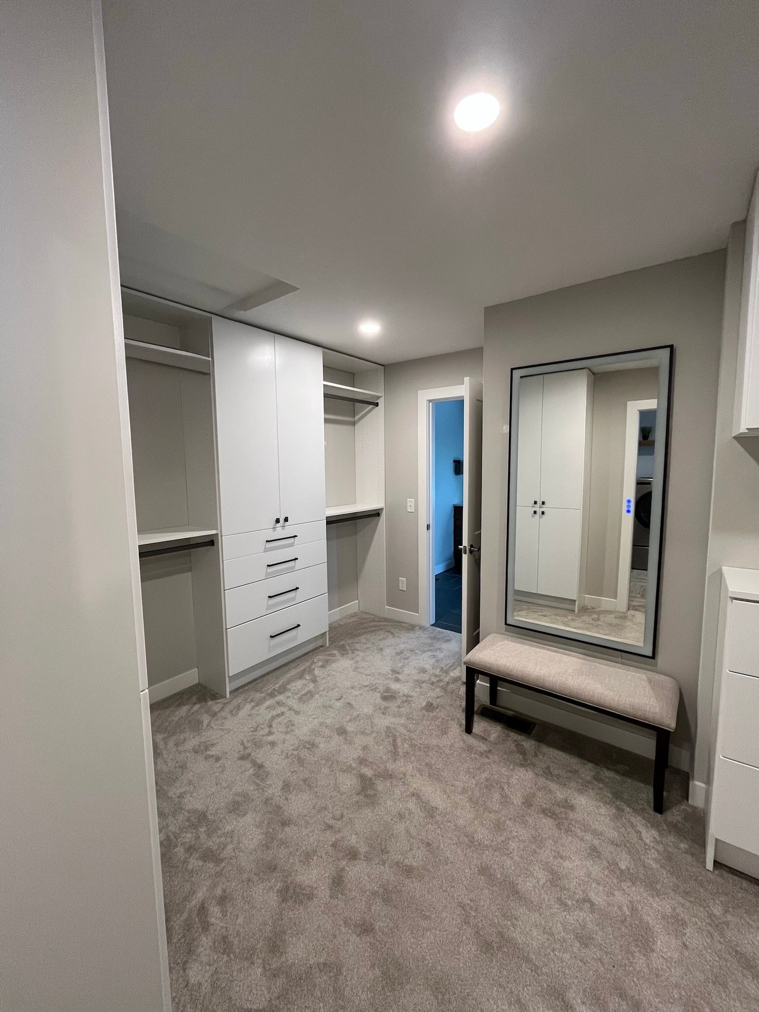 Walk-in closet with white cabinets, gray carpet, and a bench in front of a full-length mirror.