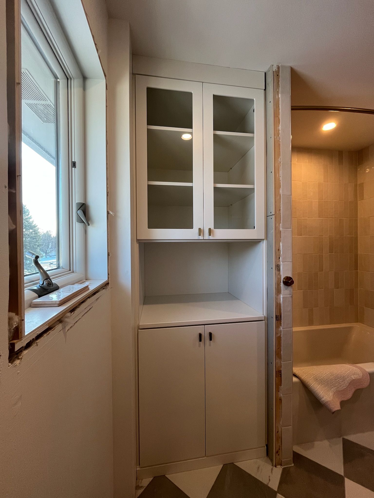 Bathroom vanity with white cabinets, mirror doors, and a window beside a tiled shower.