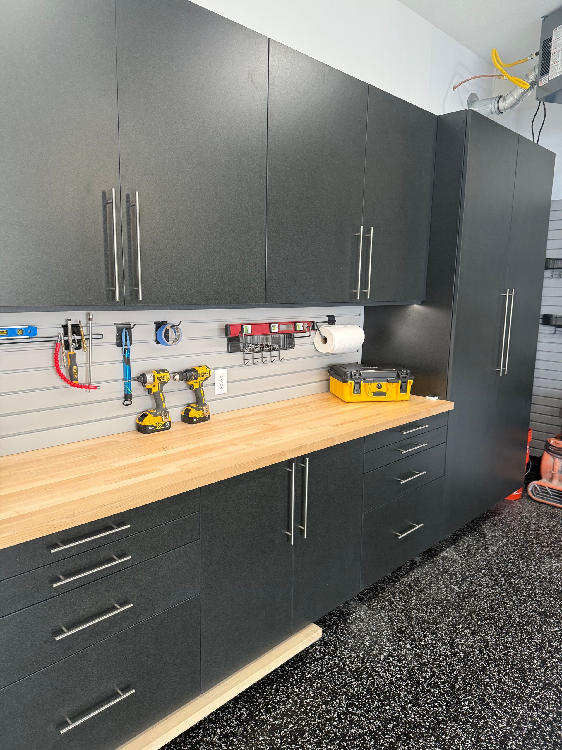 Black garage cabinets and drawers above and below a wooden workbench with tools on a speckled floor.