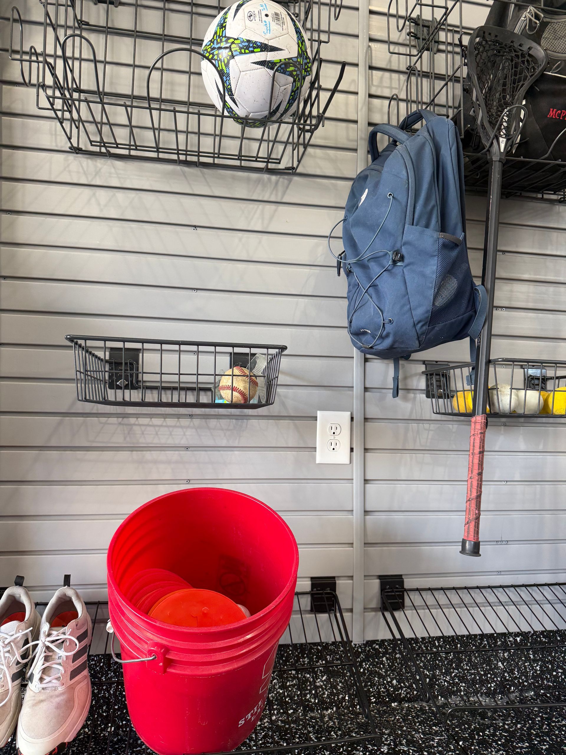 Garage wall with wire shelves holding a soccer ball, blue backpack, red buckets, and a tennis racket