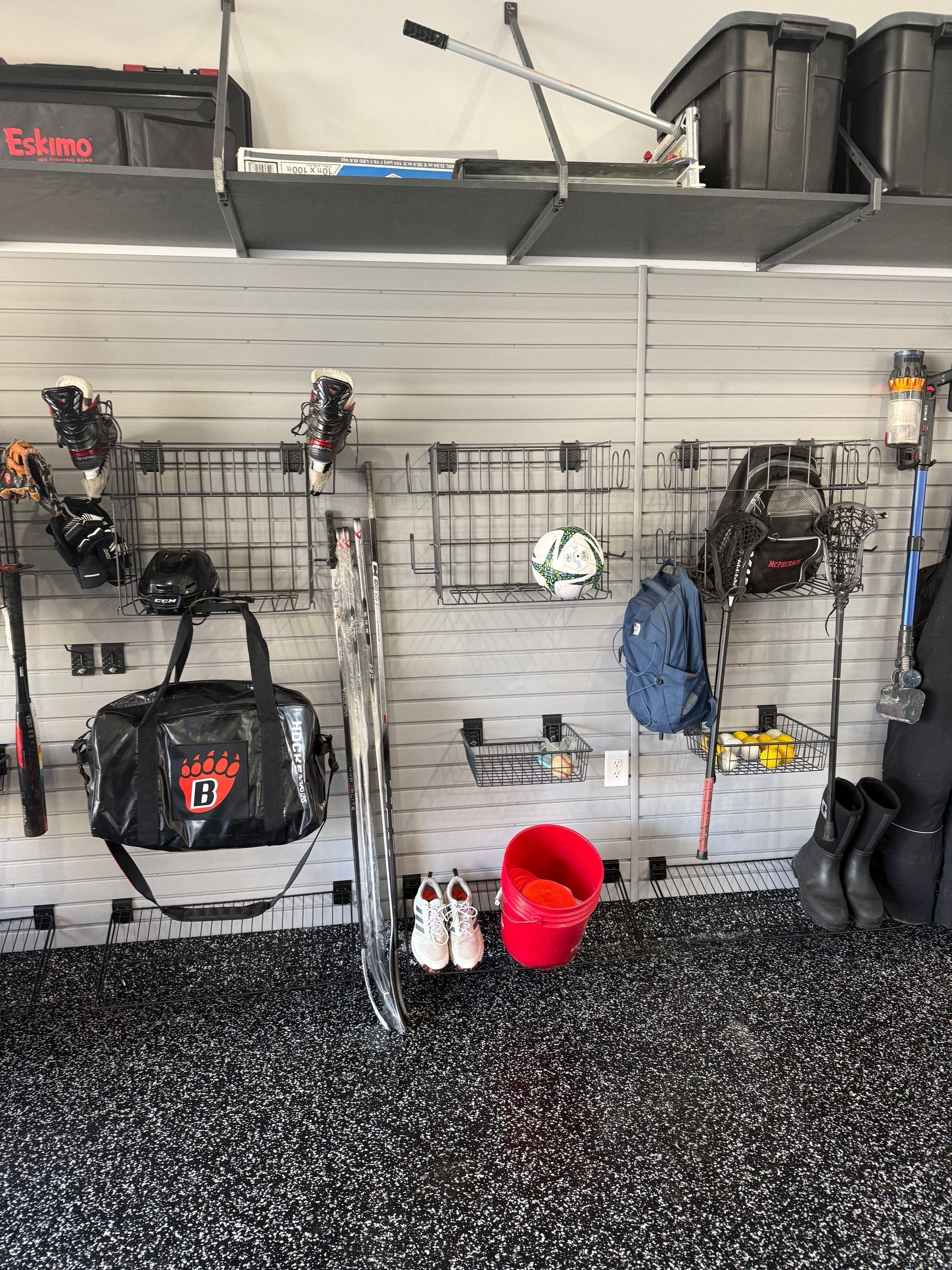 Garage wall with hanging sports gear, rackets, bins, and a red bucket on a speckled floor.