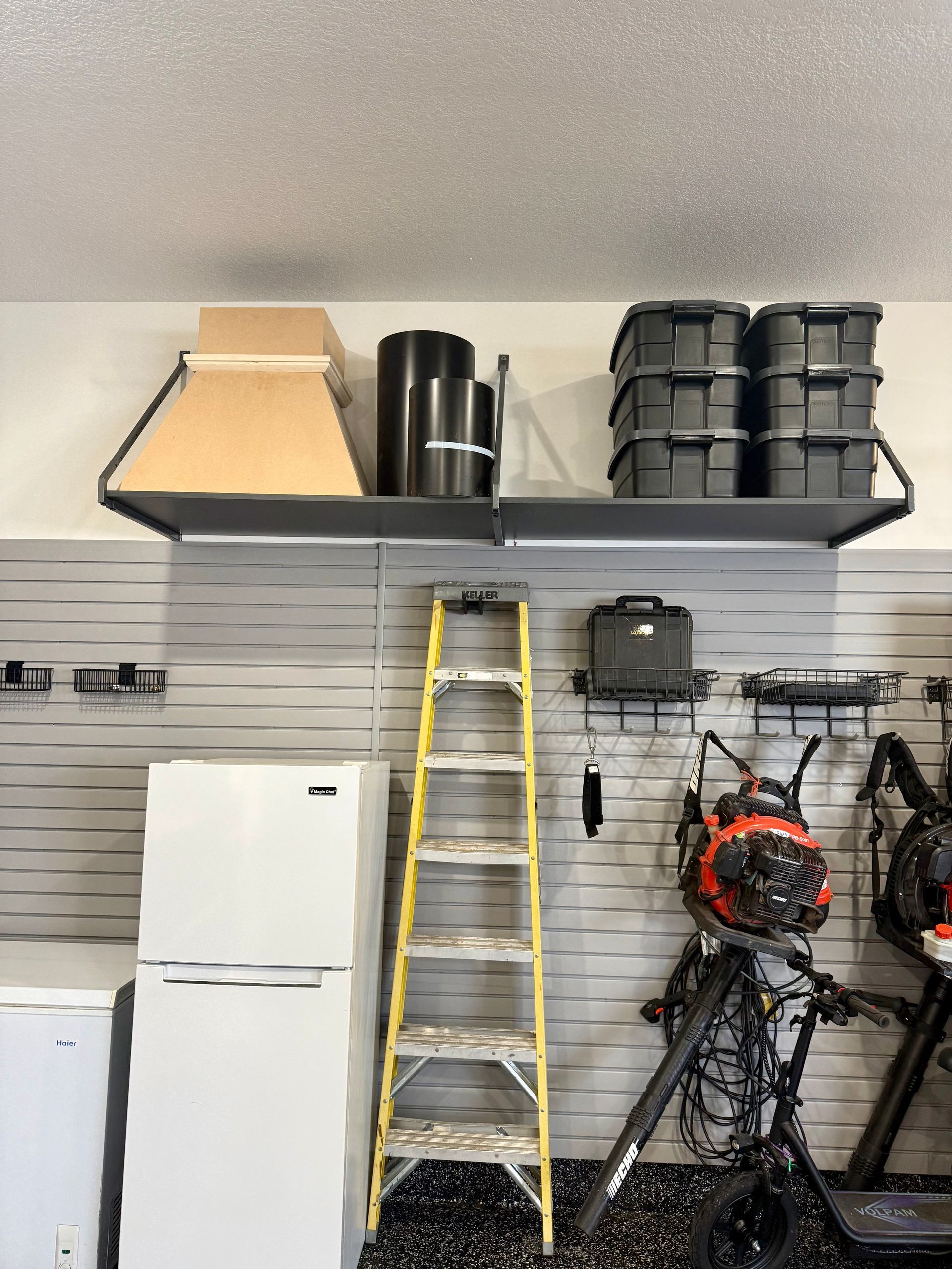 Garage wall with loft shelf, yellow ladder, white refrigerator, and hanging tools/equipment.