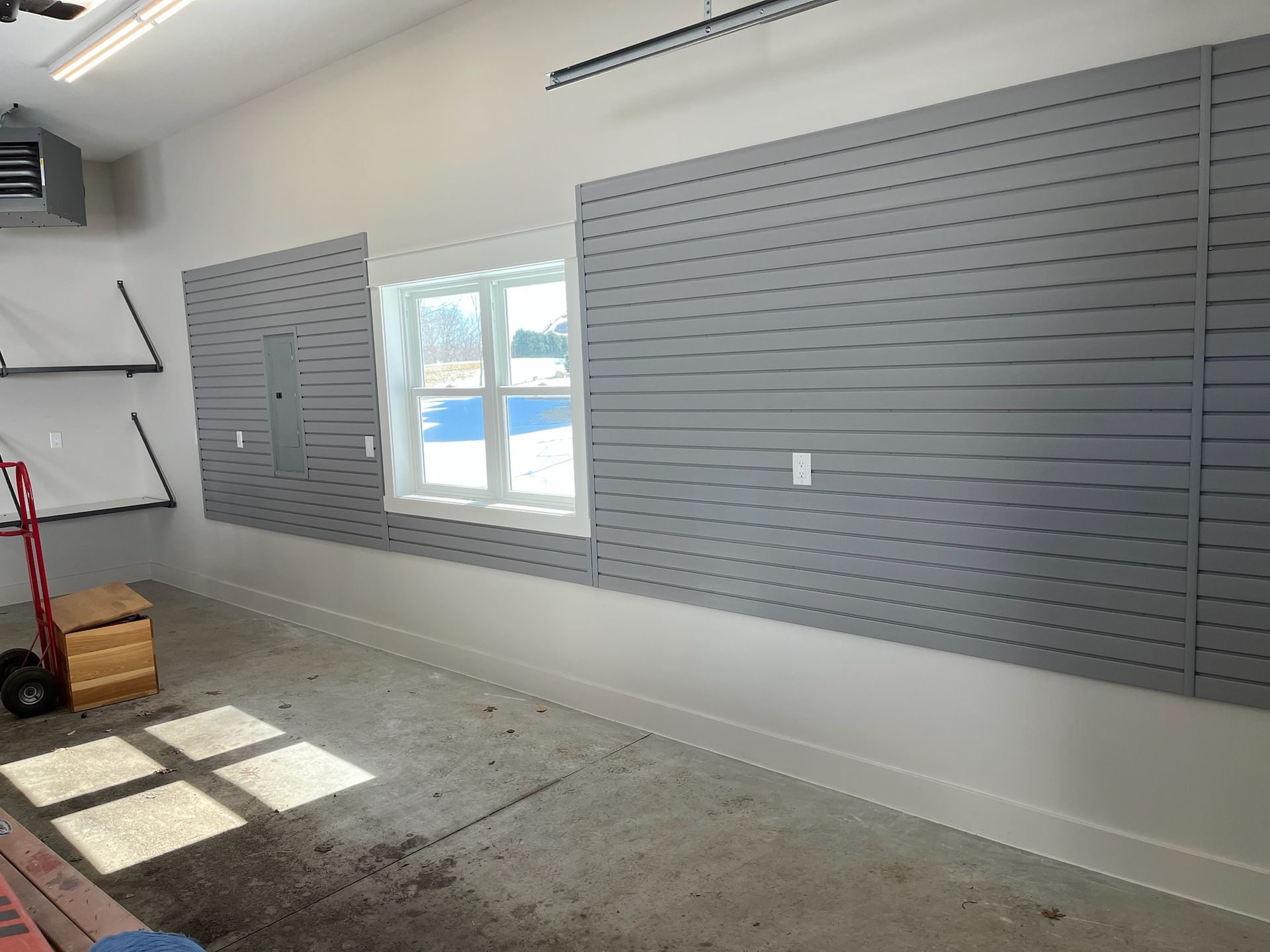 Empty garage wall with gray slatwall panels, a window, and sunlight on the floor.