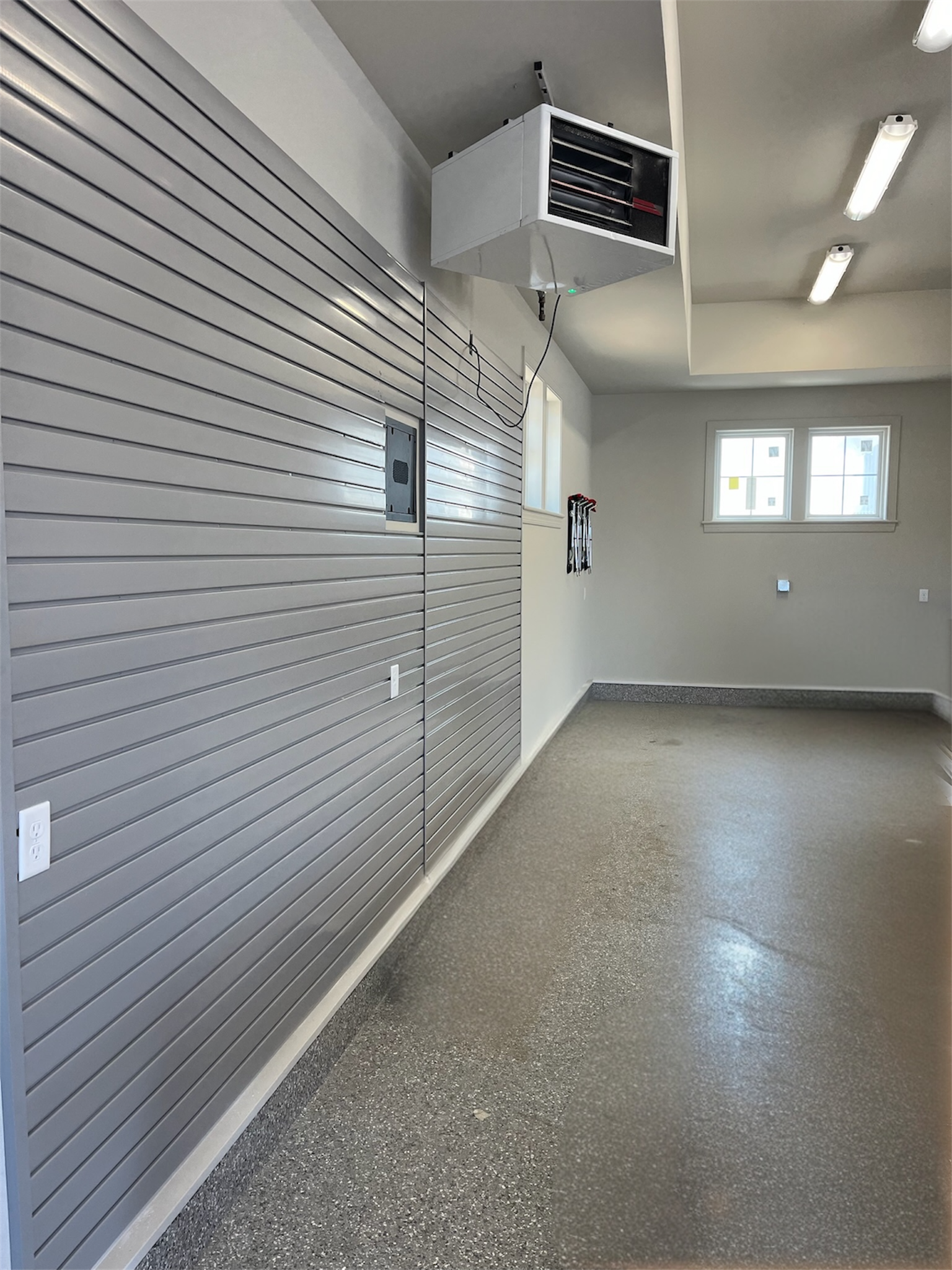 Empty garage with gray walls, slatwall storage, overhead heater, and a small window.