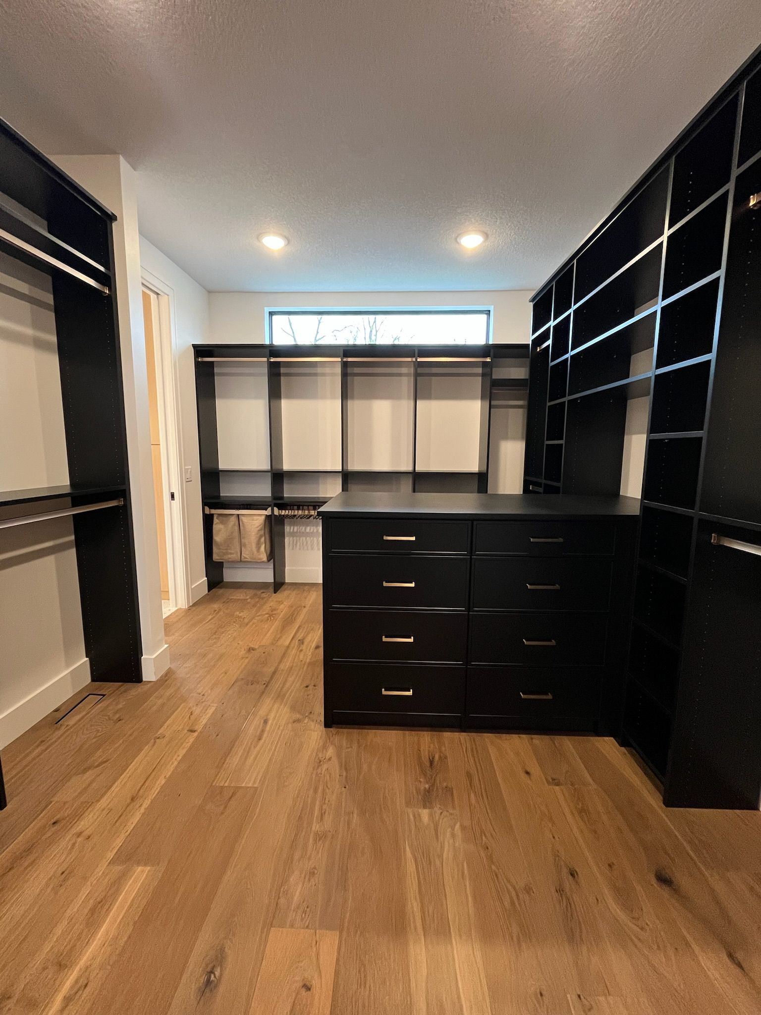 Walk-in closet with black shelving, white walls, wood floor, and a dark dresser in the center