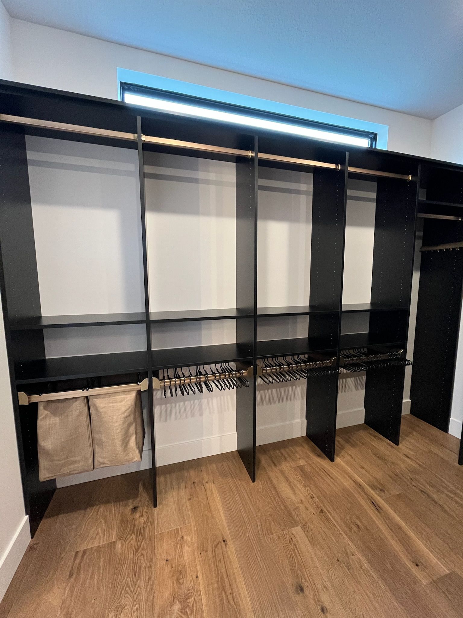 Empty walk-in closet with black shelving, hanging rods, and wood floors under a blue LED light strip