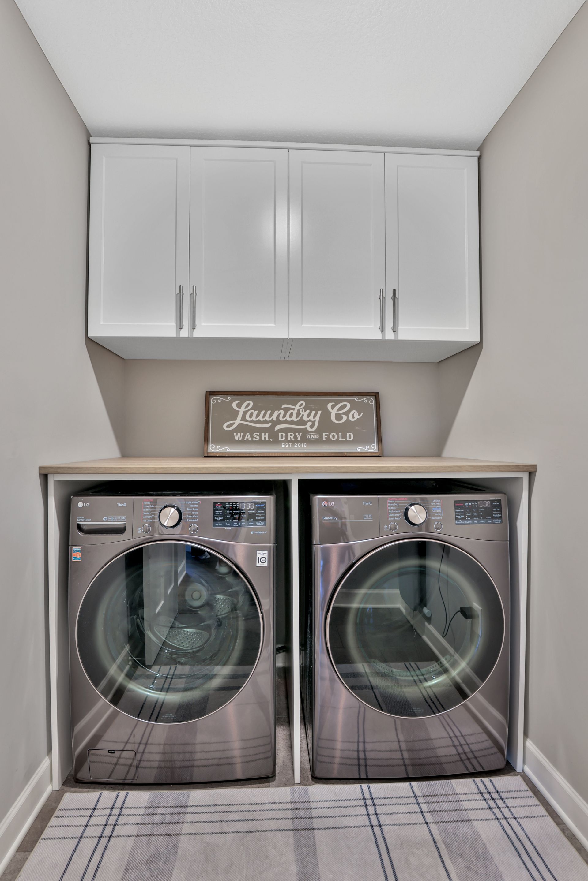 Laundry room with two front-loading washers beneath white cabinets and a decorative sign.
