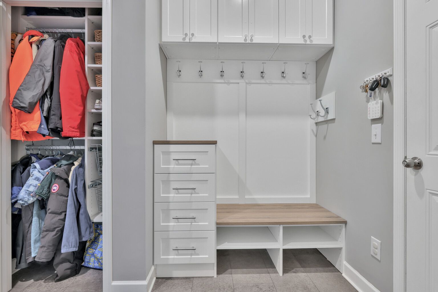 Small mudroom with white built-in bench, hooks, drawers, and an open coat closet with jackets.