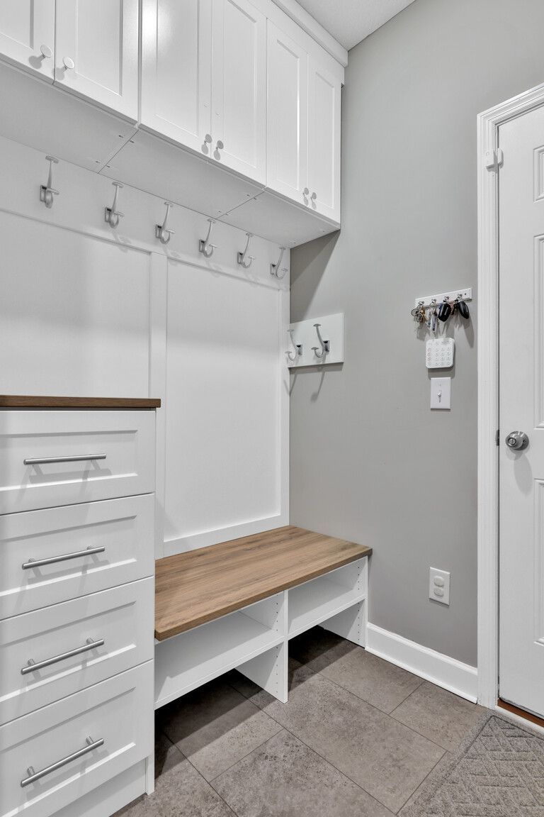 White mudroom with built-in cabinets, hooks, a wood bench, and gray tile floor.