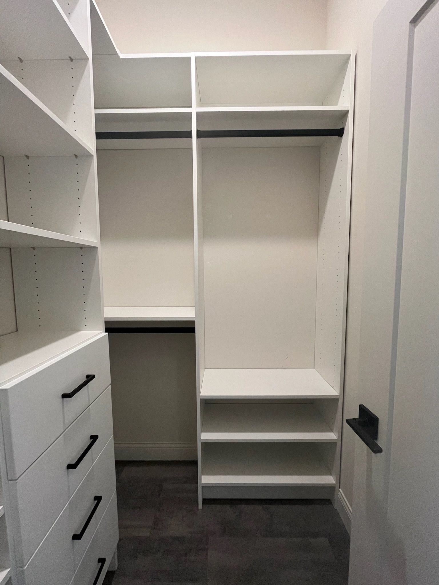 Empty walk-in closet with white shelves, drawers, hanging rods, and a black door handle.