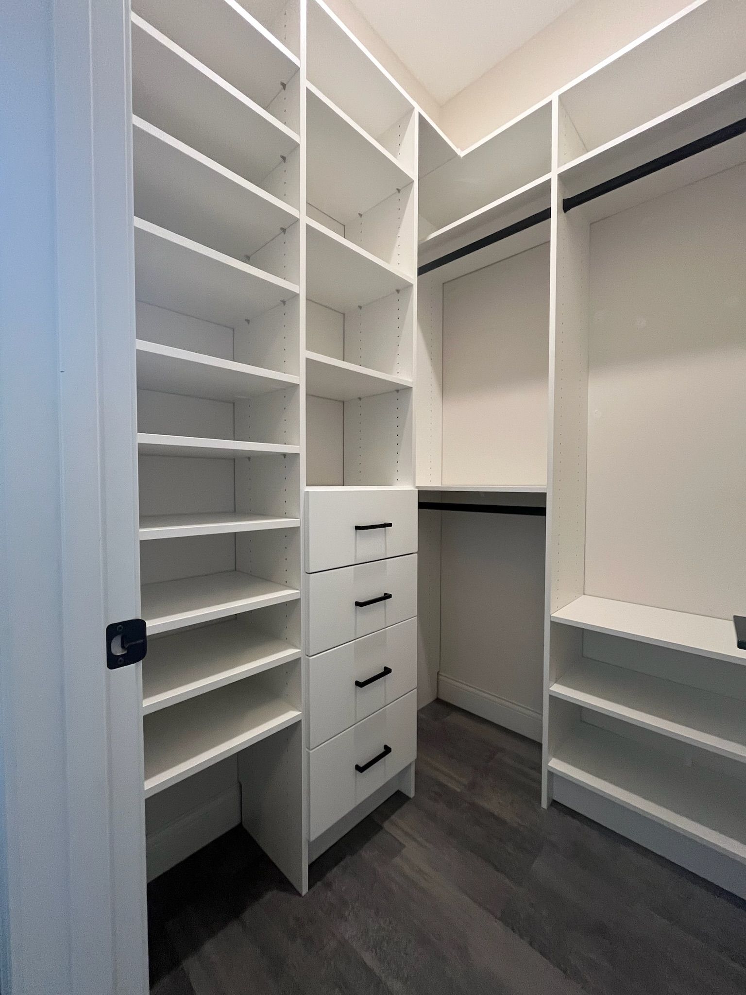 Empty white walk-in closet with shelves, drawers, and hanging rods on dark flooring.
