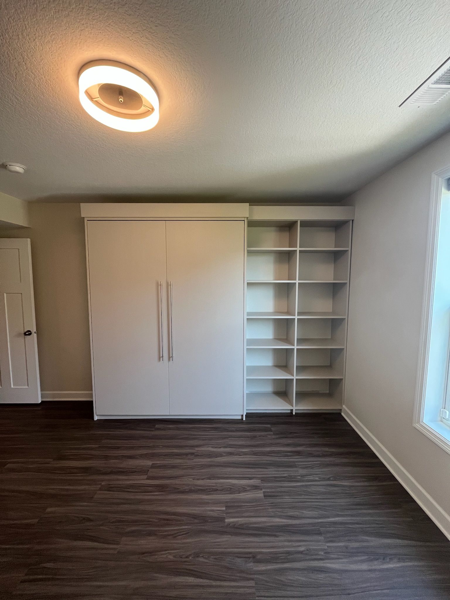 Empty bedroom with white cabinet and shelves, dark wood floor, ceiling light, and window blinds on the right.