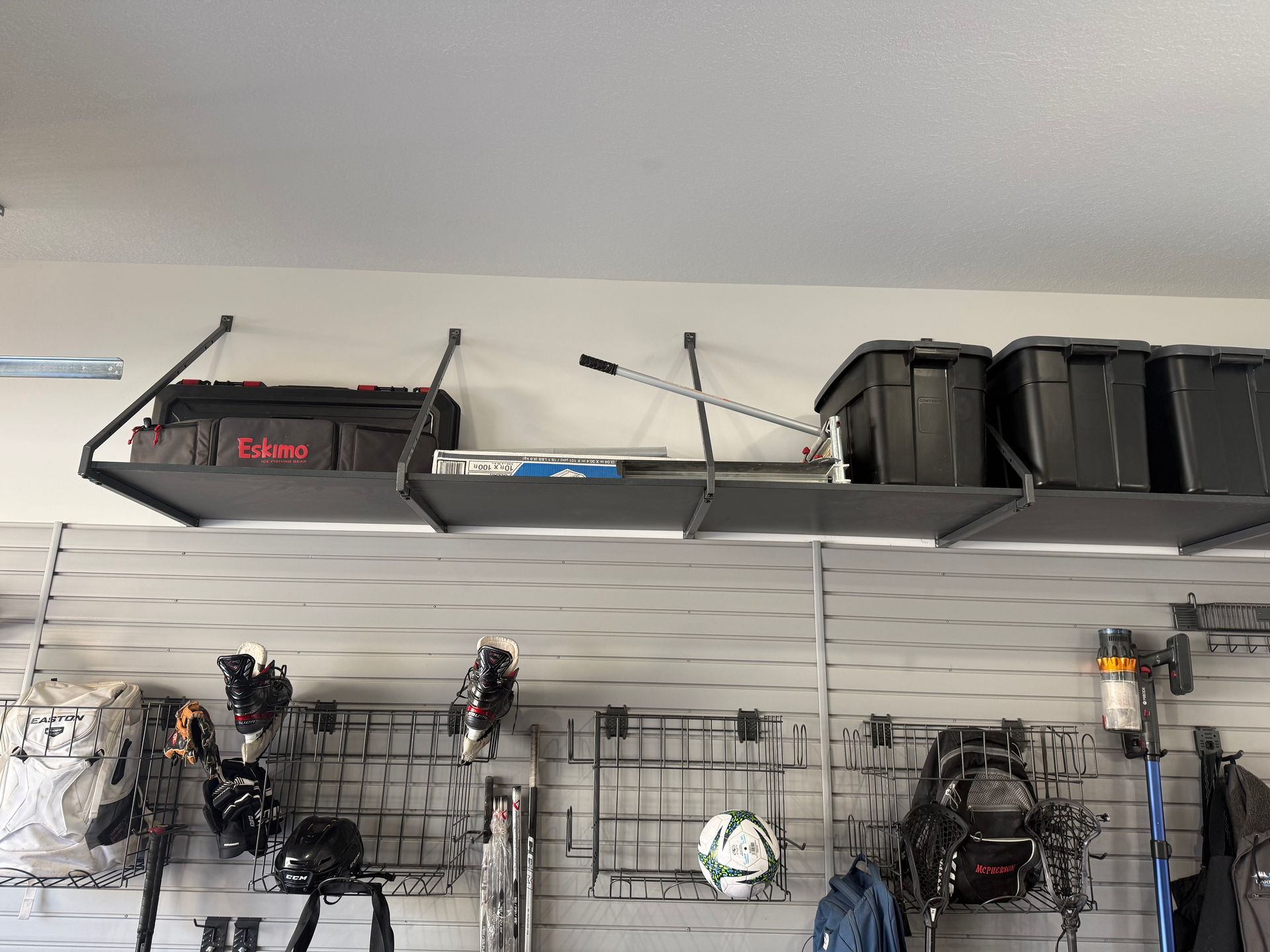 Garage wall with storage shelves holding bins and equipment, and several tools hanging on a slat wall.