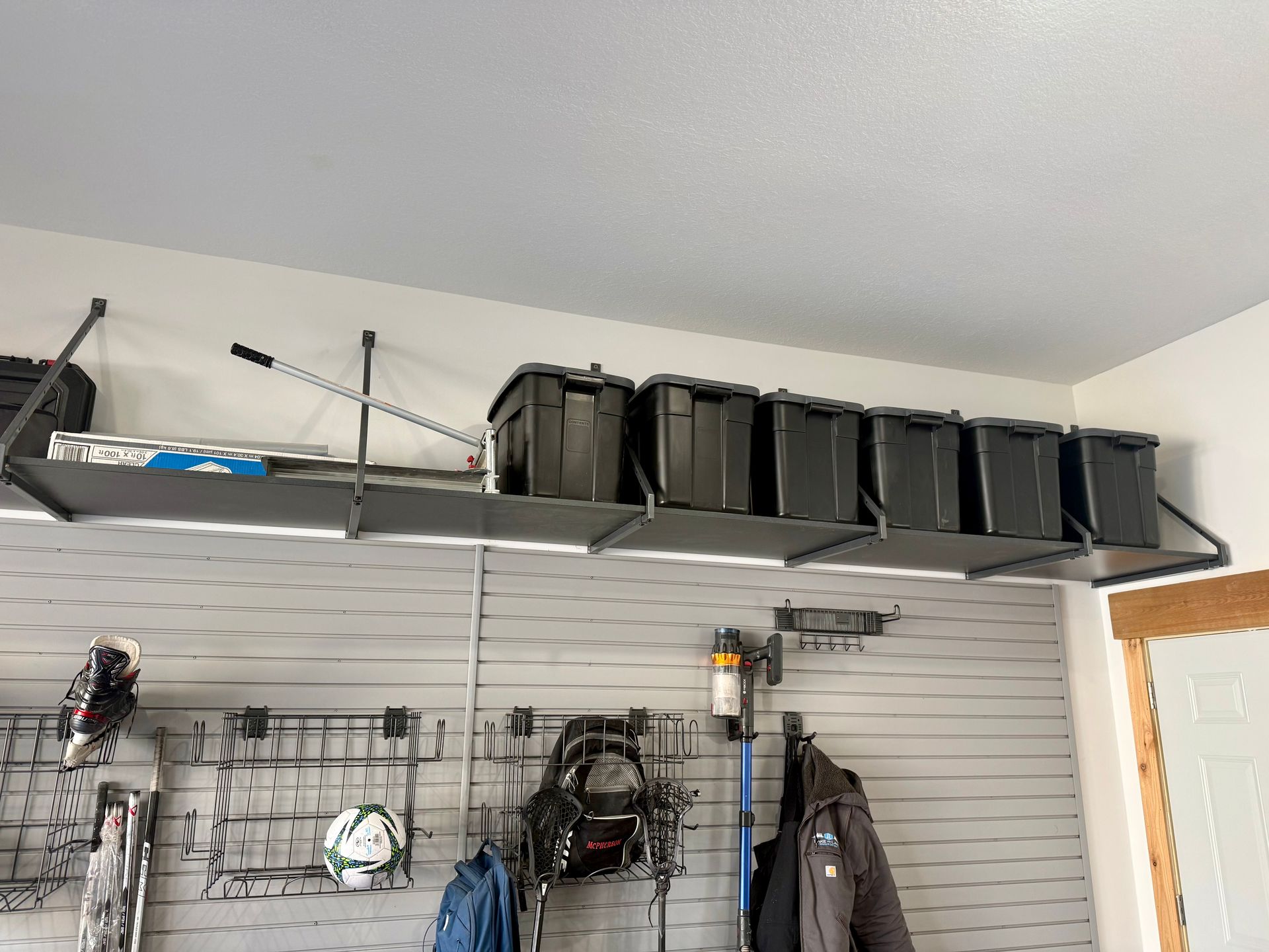 Garage wall shelves with black storage bins, fishing rods, and hanging gear above a slatwall organizer