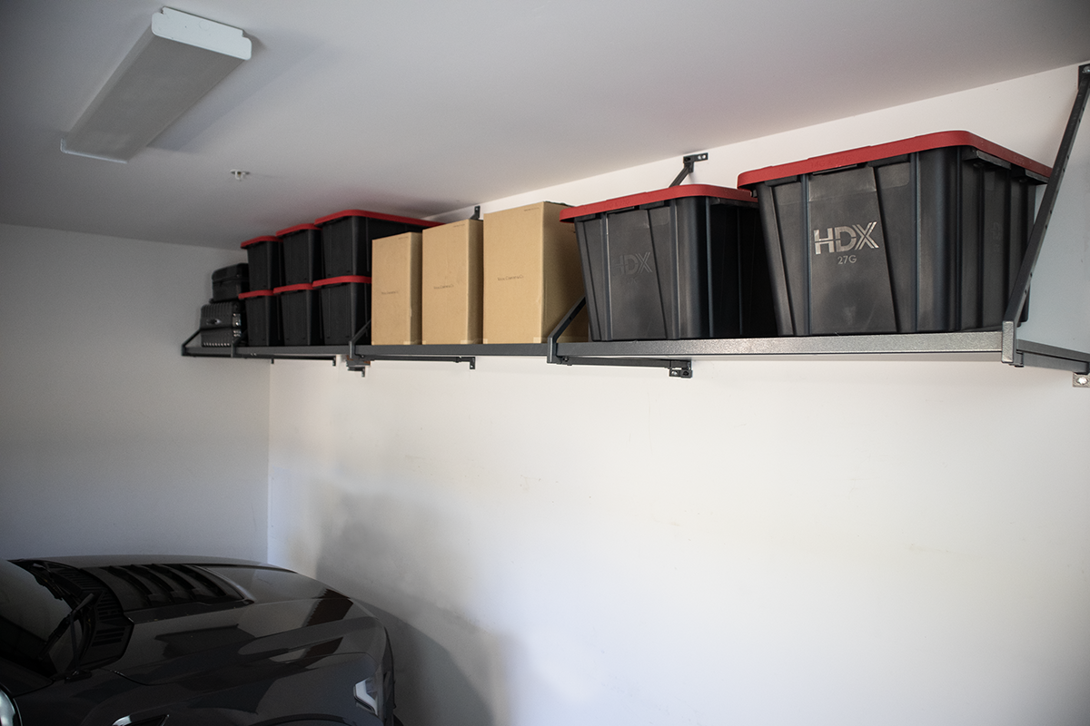Overhead garage storage shelf holding black bins and cardboard boxes.
