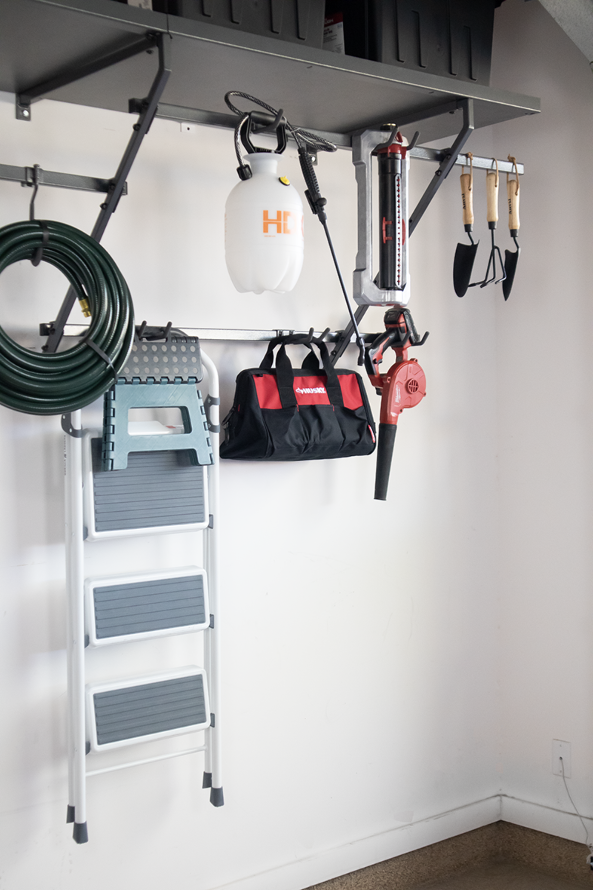 Garage wall storage with ladder, hose, tools, sprayer, and tote bag.