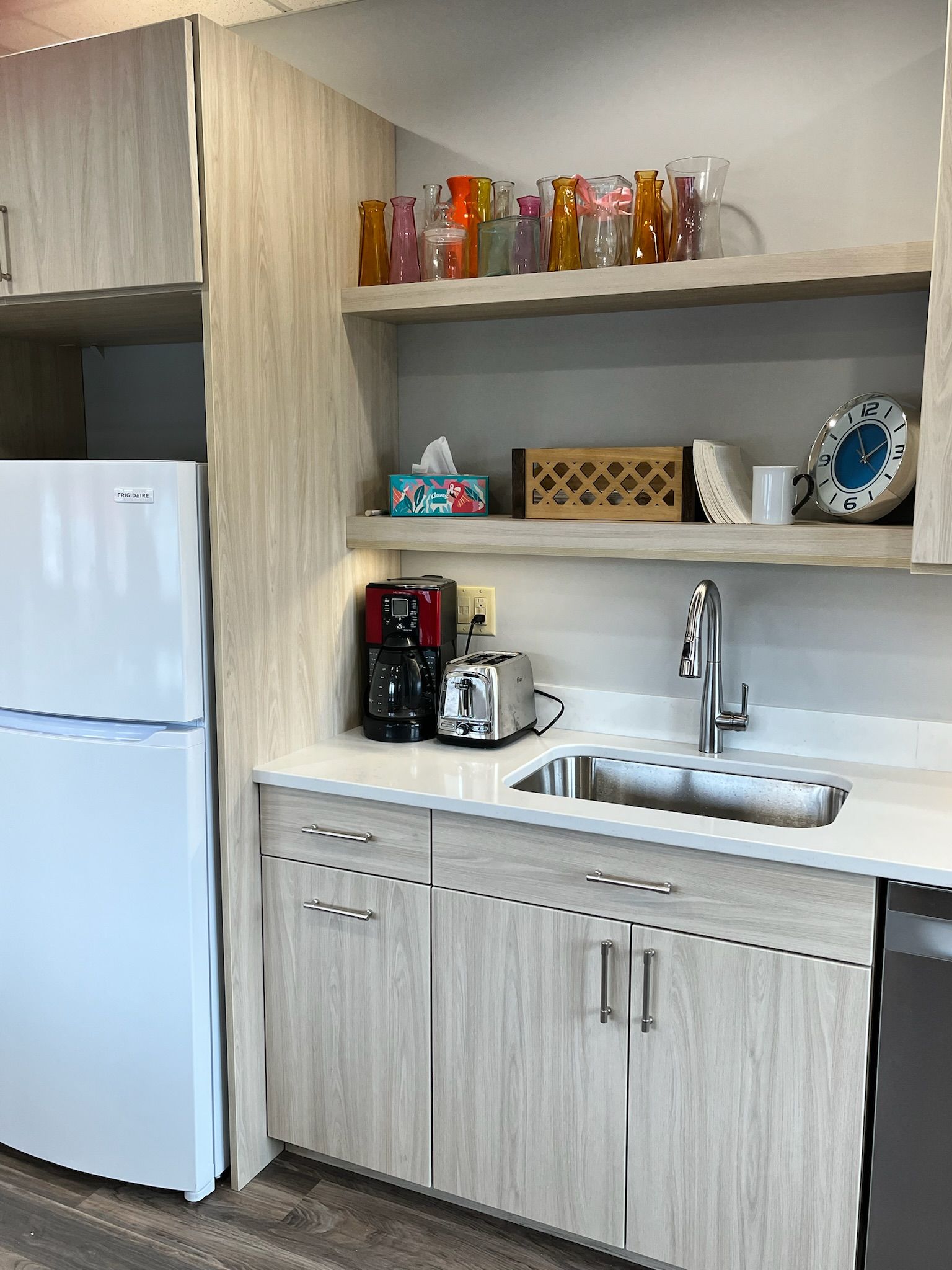Small kitchenette with white fridge, light wood cabinets, sink, and shelves holding mugs and snacks.