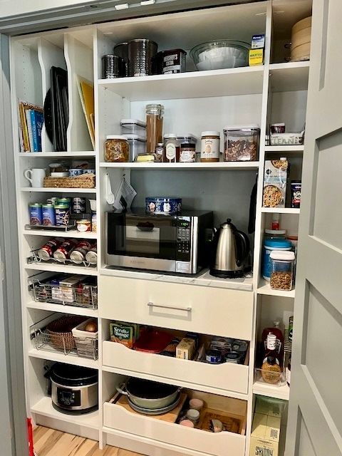 White pantry cabinet with microwave, kettle, jars, spices, mugs, and food containers organized on shelves.