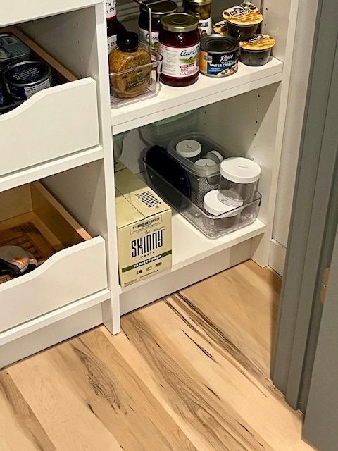 Open pantry cabinet with jars, canned goods, and a box on white shelves beside a wood floor.