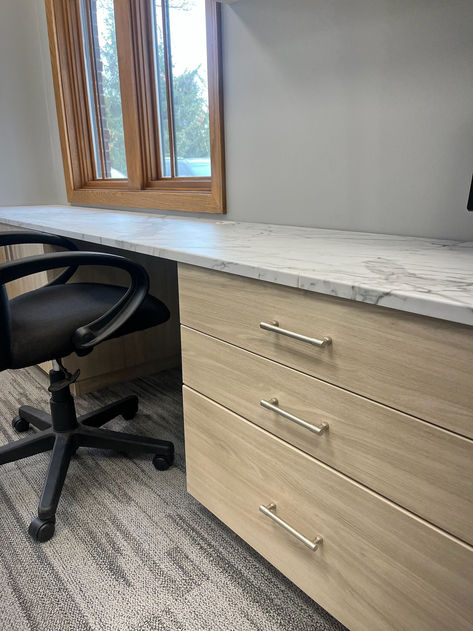 Office desk with light wood drawers, white countertop, black chair, and window