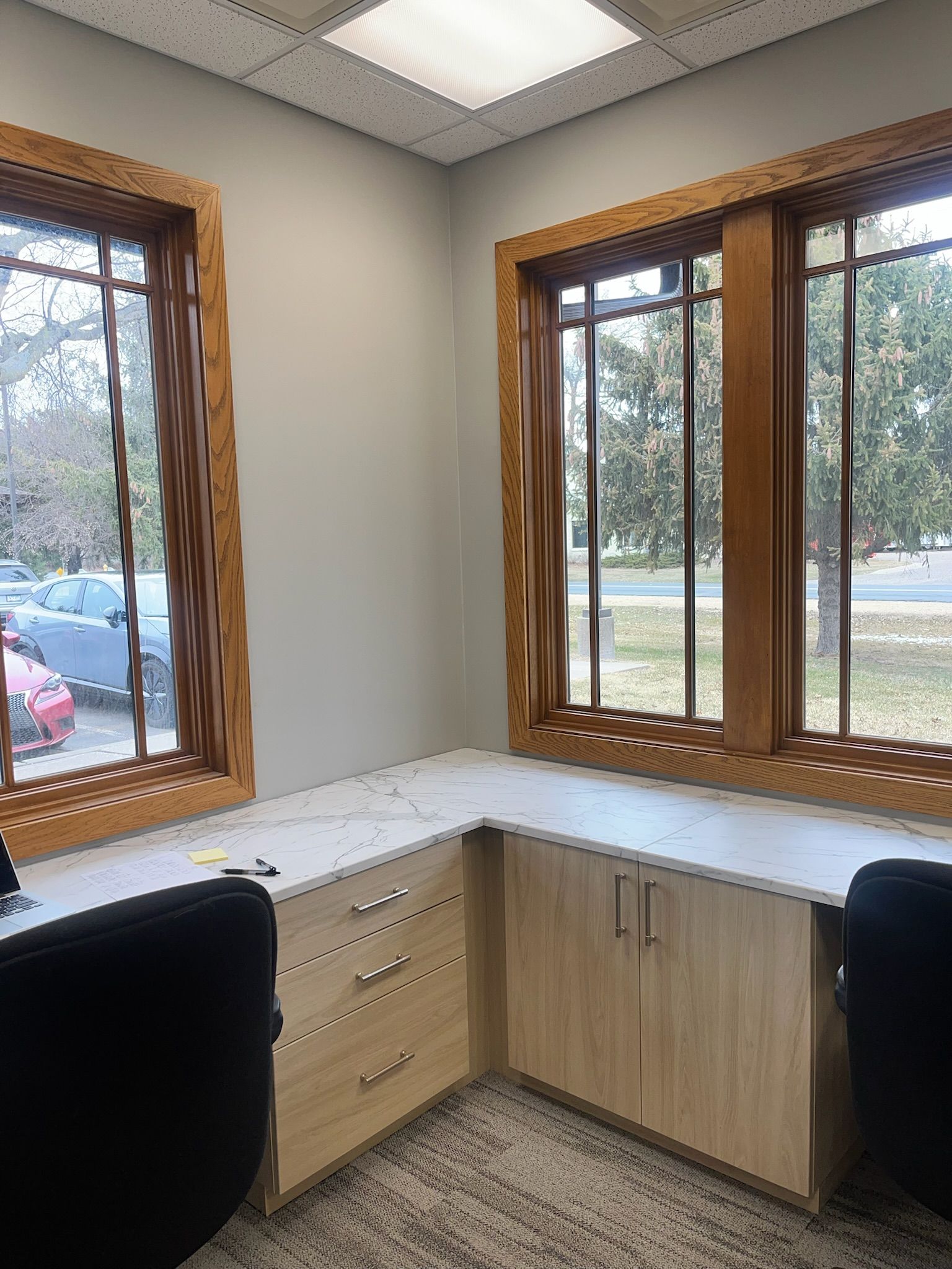 Empty office corner with desk, cabinet, chair, and large windows overlooking trees and parked cars