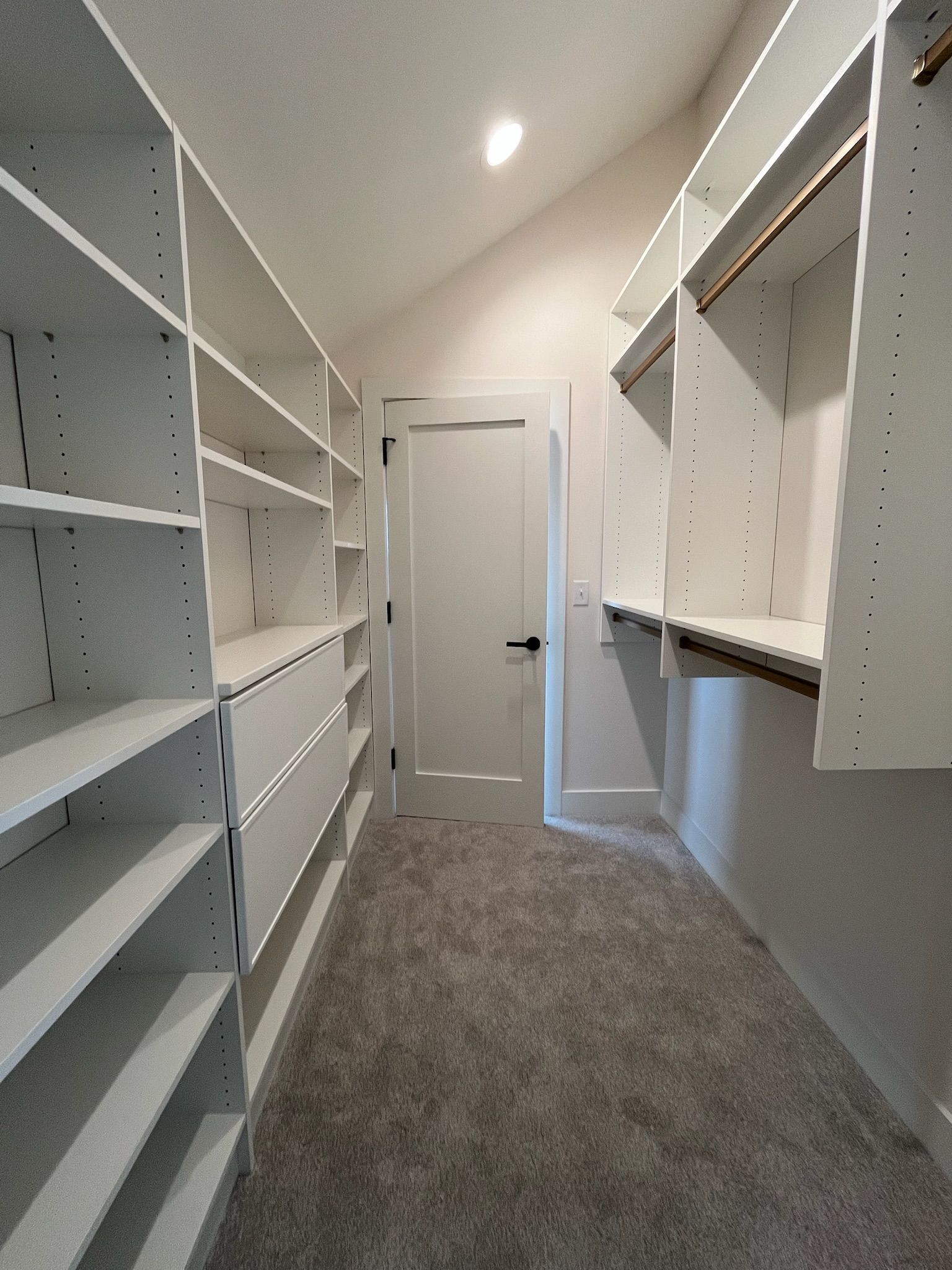 Narrow walk-in closet with white shelves, hanging rods, gray carpet, and a door at the end