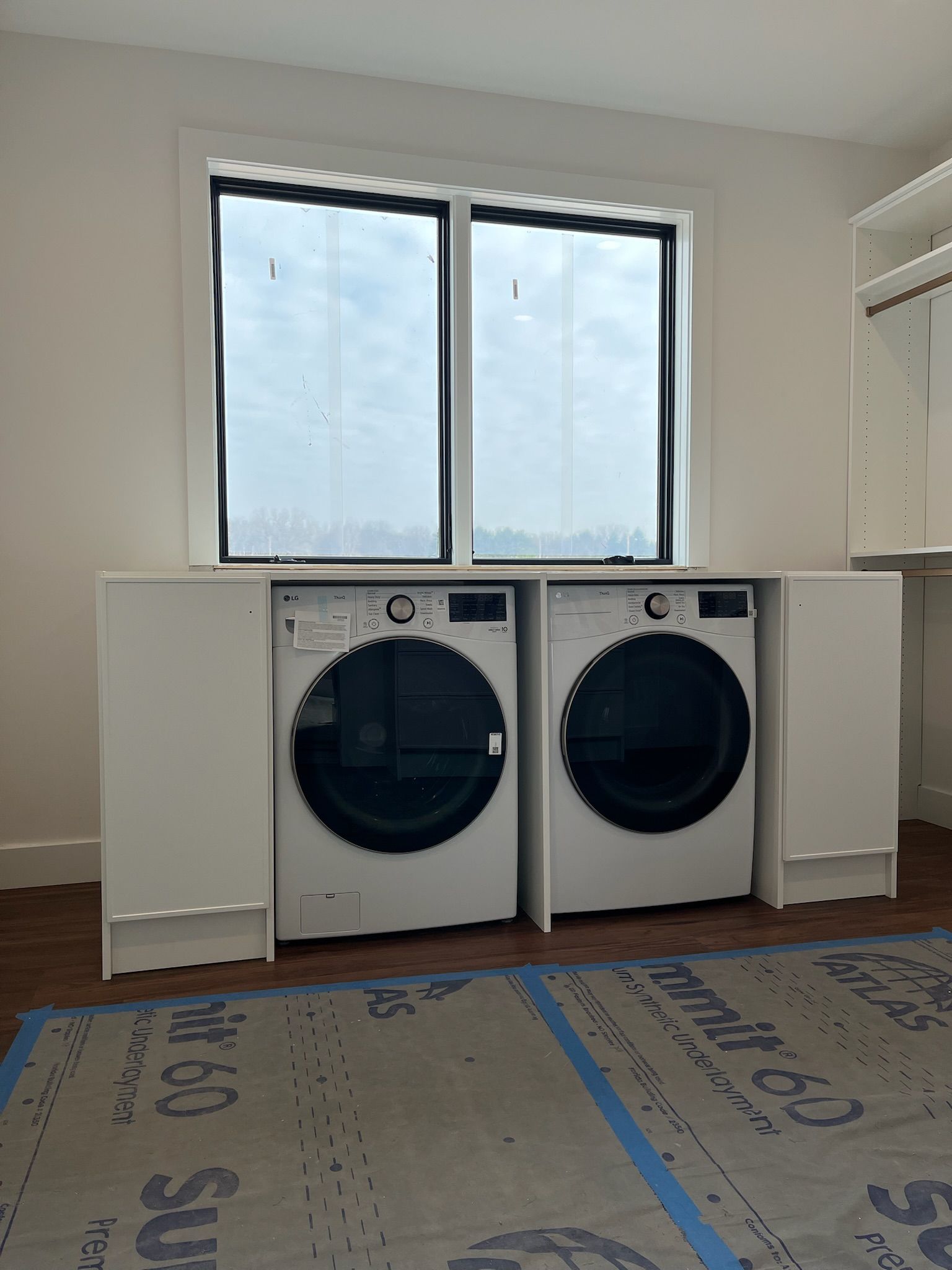 Laundry room with two front-load machines under a window, white cabinets, and unfinished flooring.