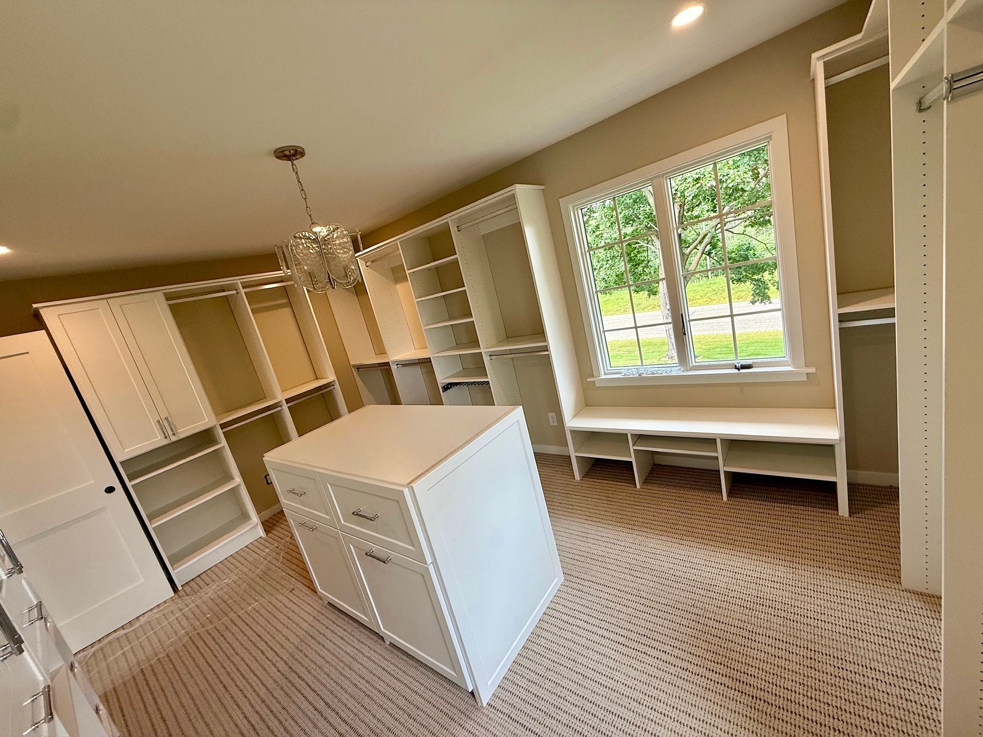 Bright walk-in closet with white shelving, central island, beige carpet, and a large window overlooking trees