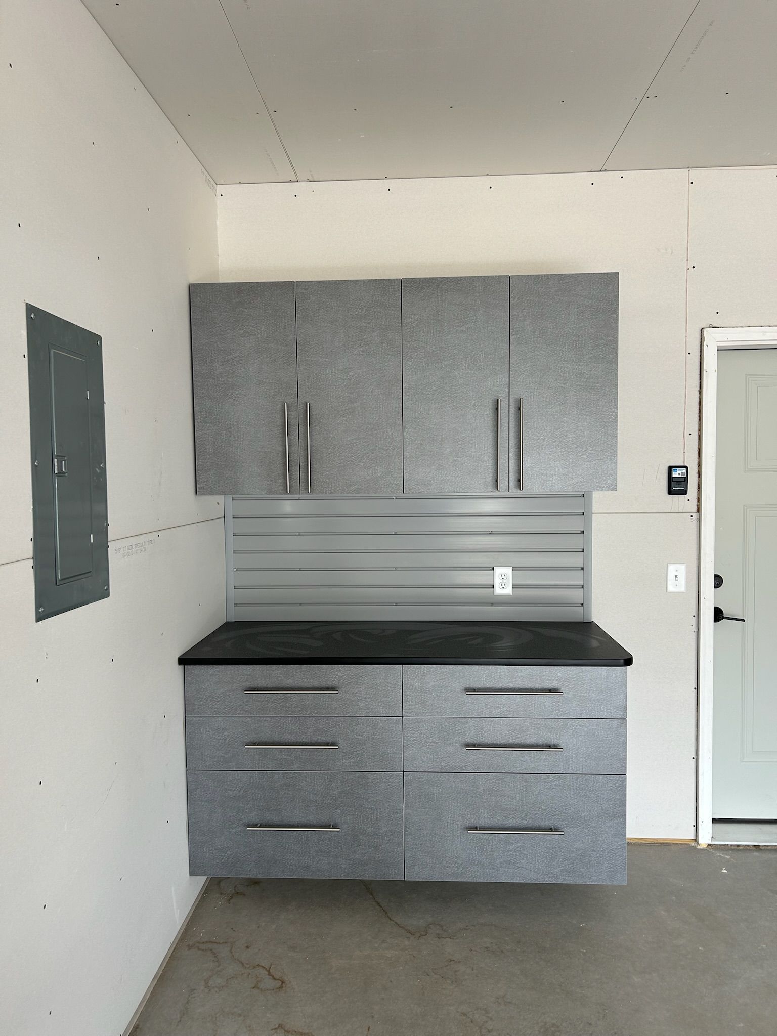 Gray wall-mounted cabinets and drawers in a garage-style room with a black countertop and utility panels