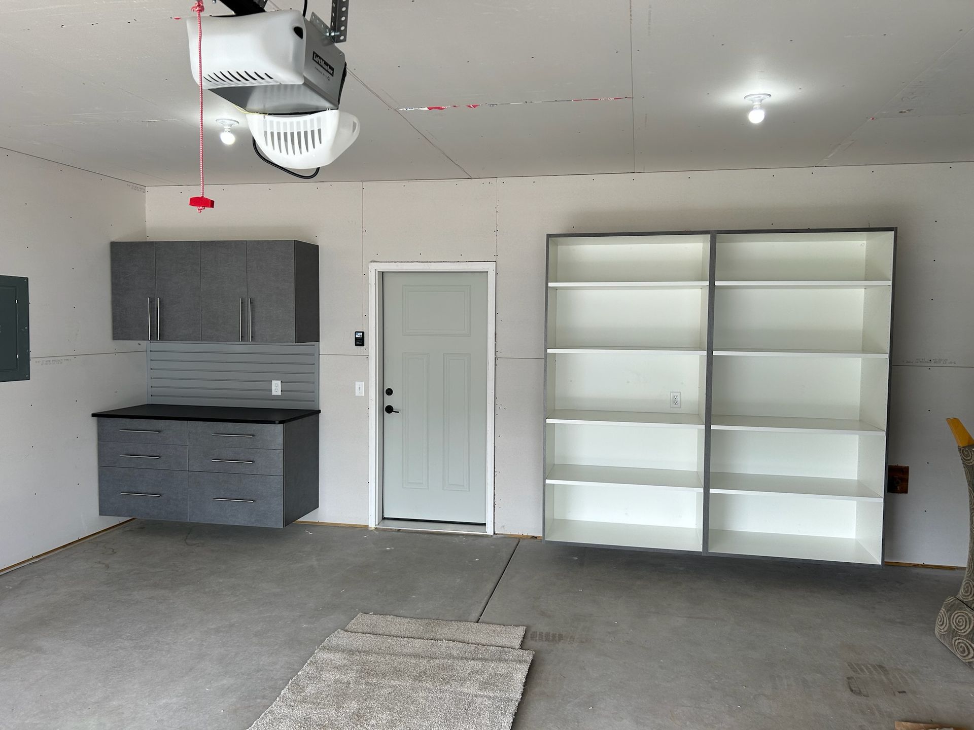 Empty garage with gray cabinets, white door, tall shelving, and a gray carpet remnant on the floor.