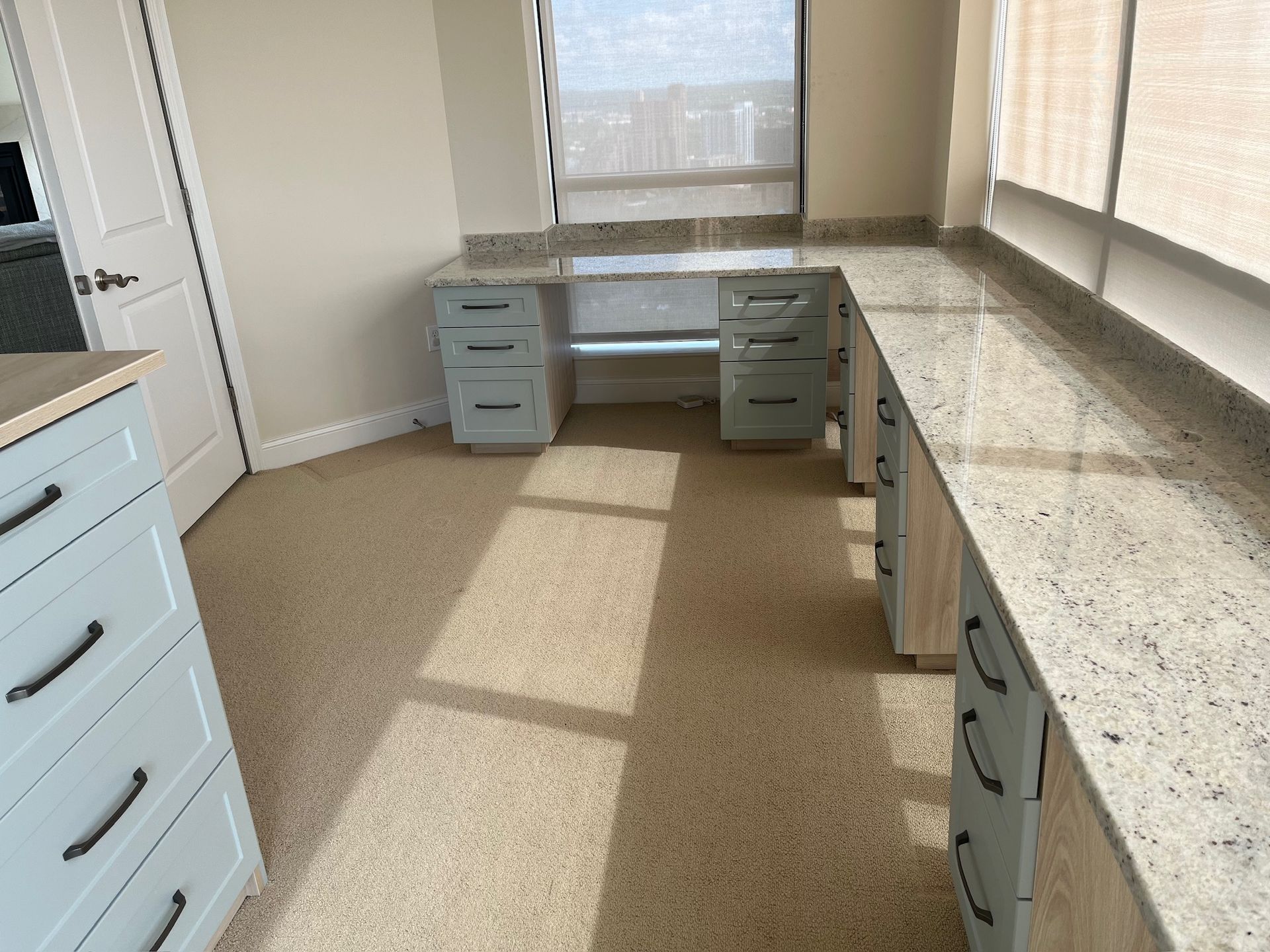 Empty office with blue filing drawers, built-in desk, and sunlit carpeted floor by windows