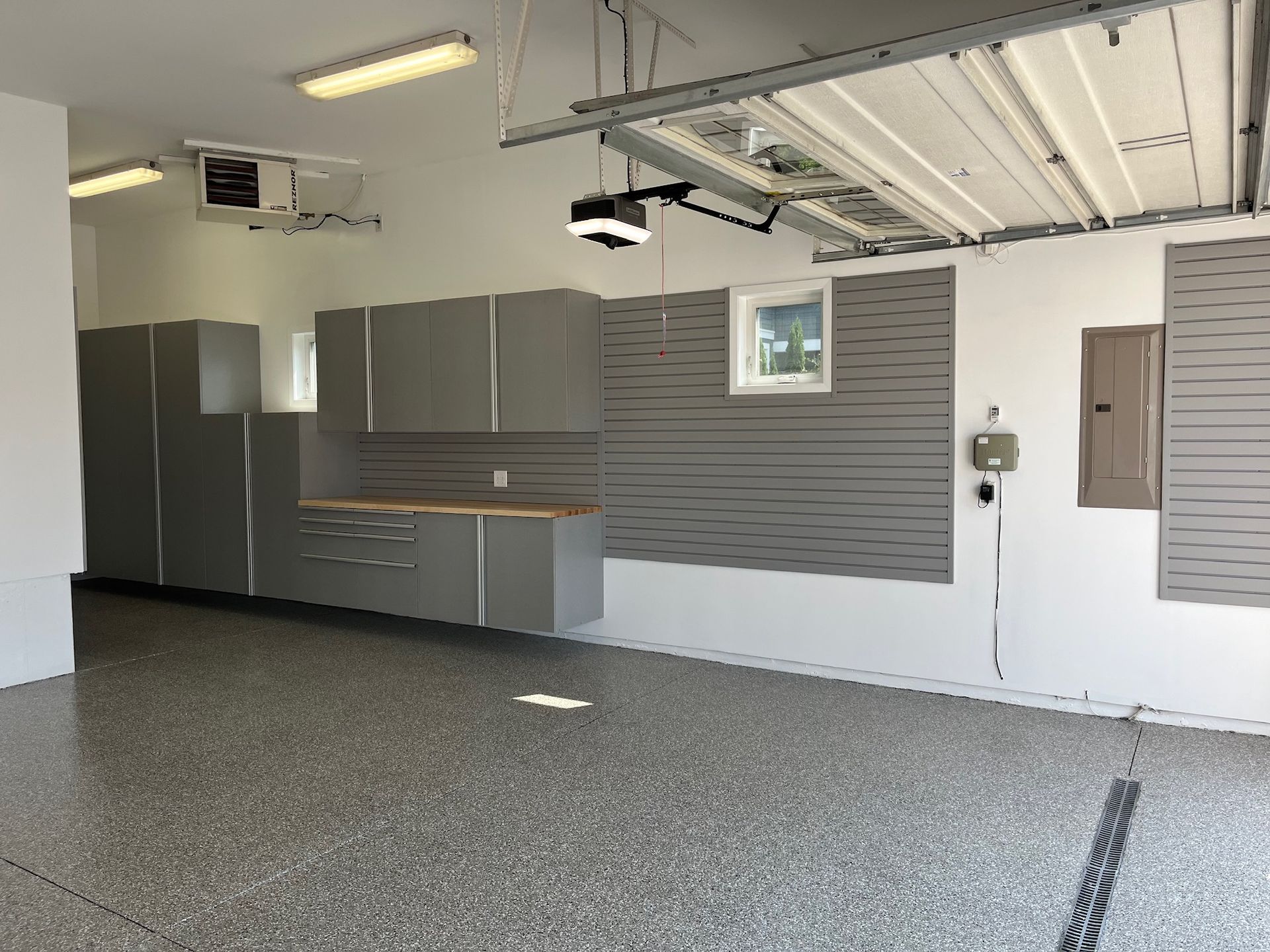 Empty garage interior with gray cabinets, white walls, and a speckled floor under fluorescent lights