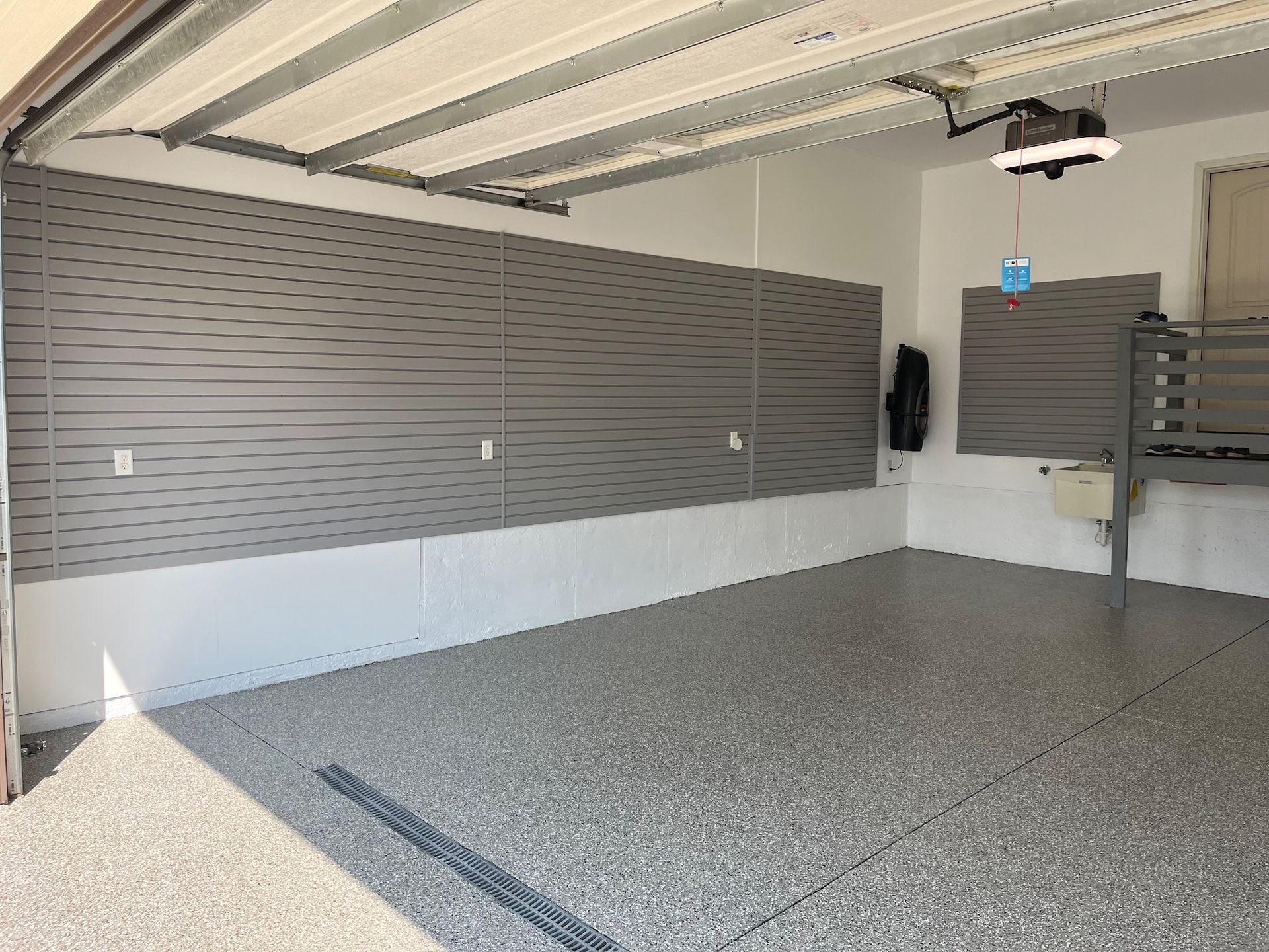 Empty garage with gray slatwall panels and speckled concrete floor, open overhead door letting in sunlight